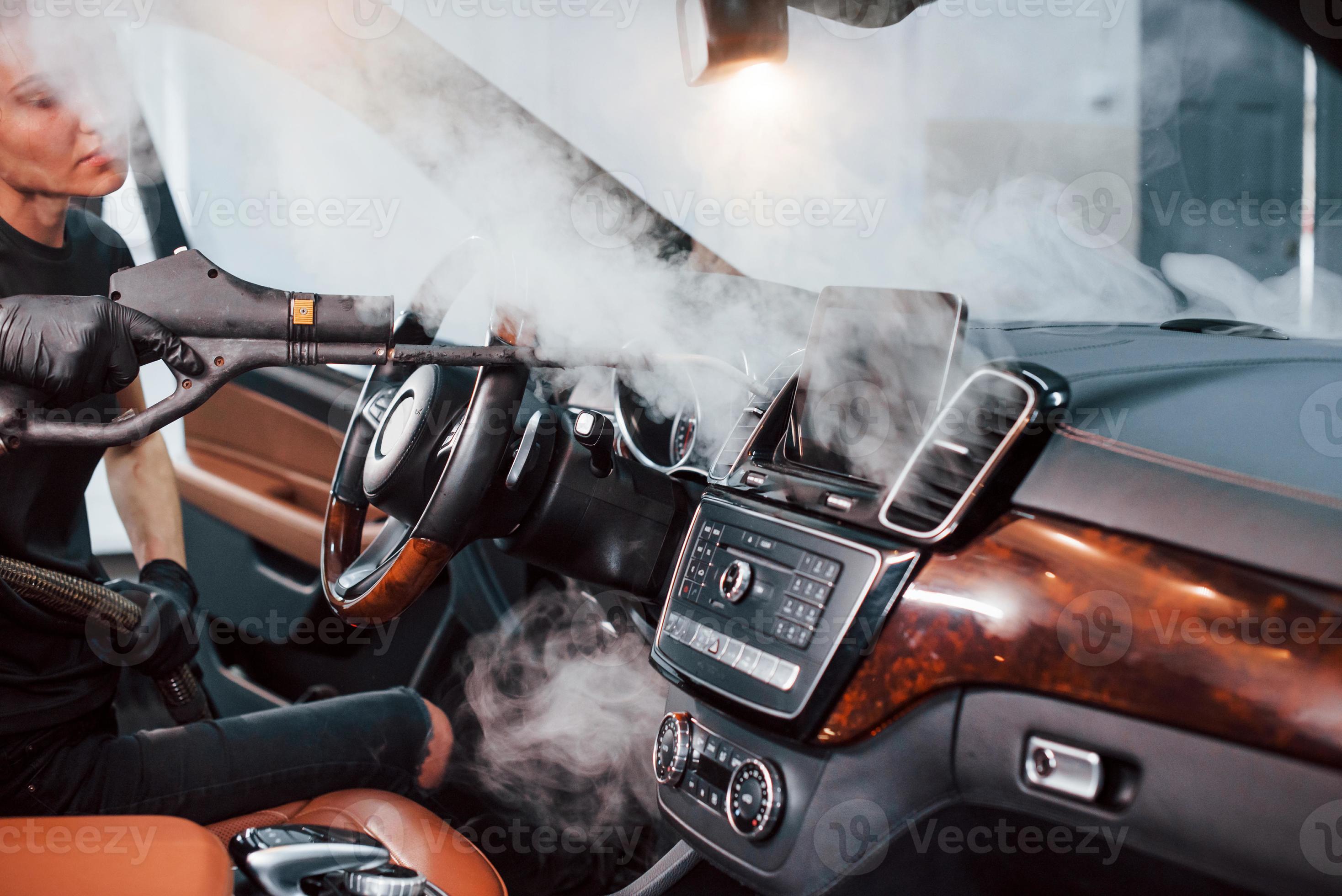 Modern black automobile get cleaned inside by steam equipment by worker inside of car wash