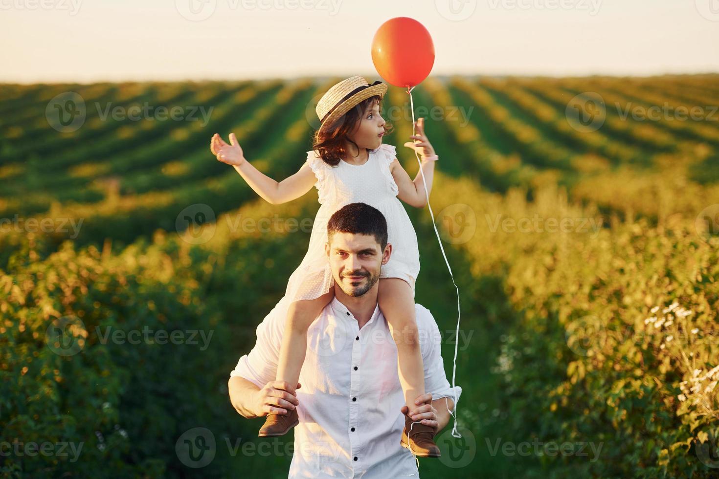 Father with his daughter that sits on shoulders and holds red balloon spending free time on the field at sunny day time of summer photo