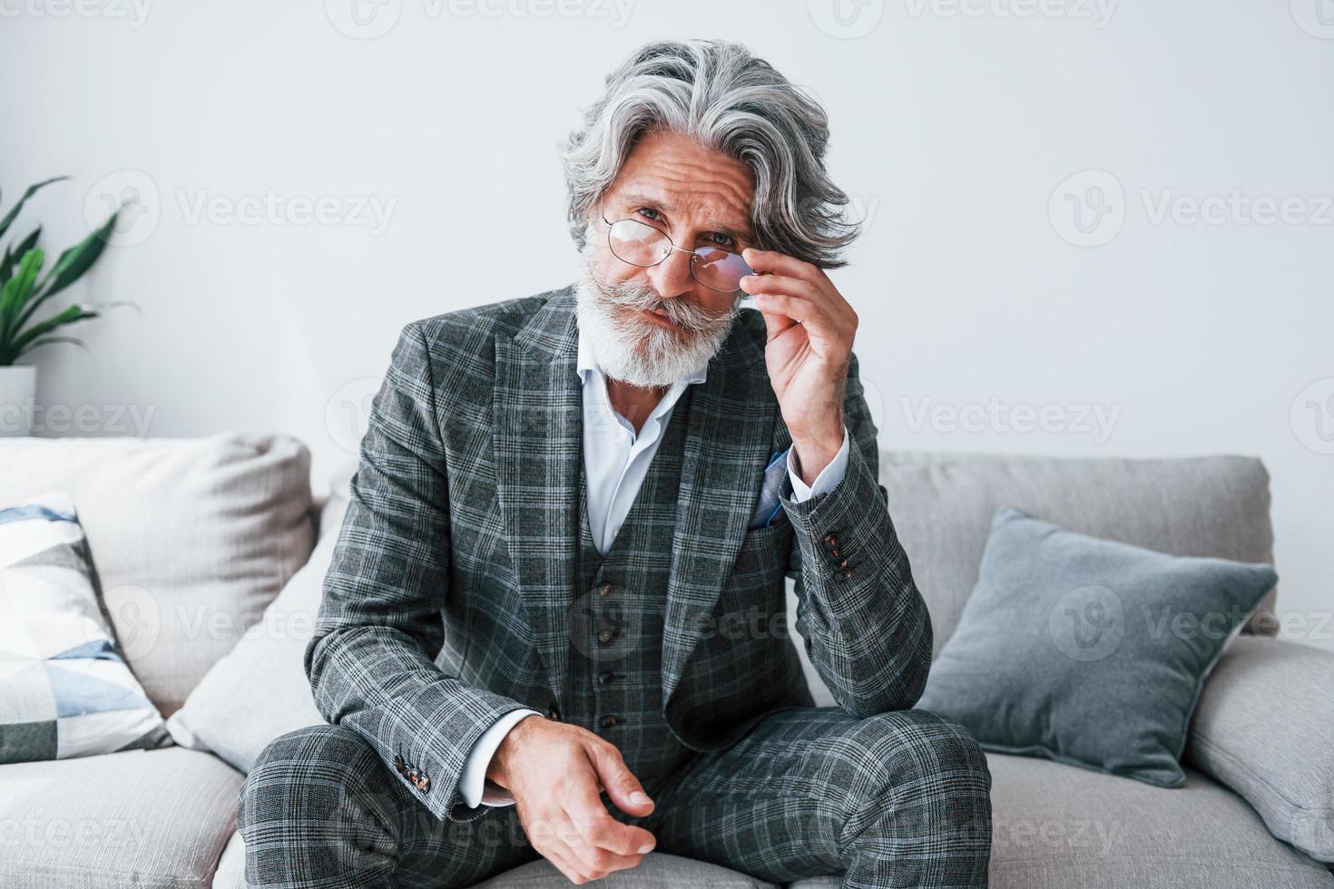 Sits on comfortable sofa in formal clothes. Senior stylish modern man with grey hair and beard indoors photo