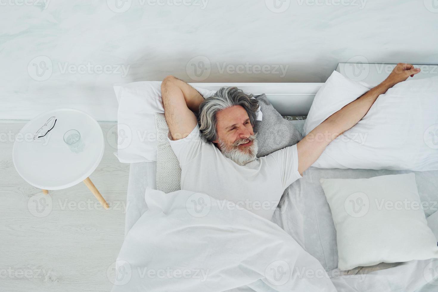 Senior stylish modern man with grey hair and beard indoors lying on the bed and waking up photo