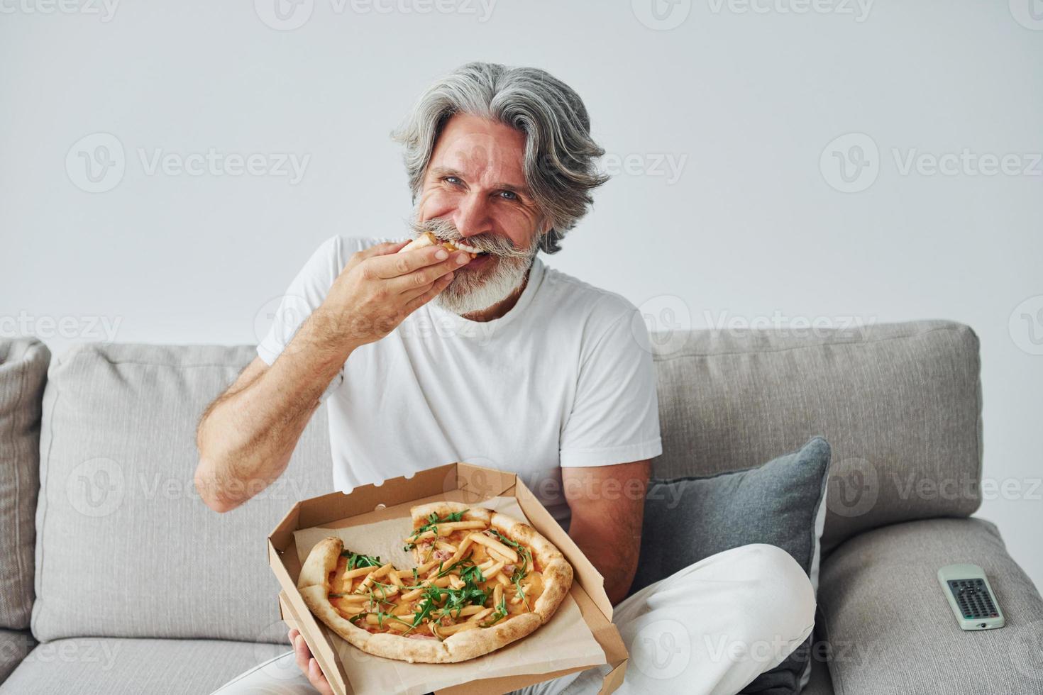 Eats delicious pizza while watching TV show. Senior stylish modern man with grey hair and beard indoors photo