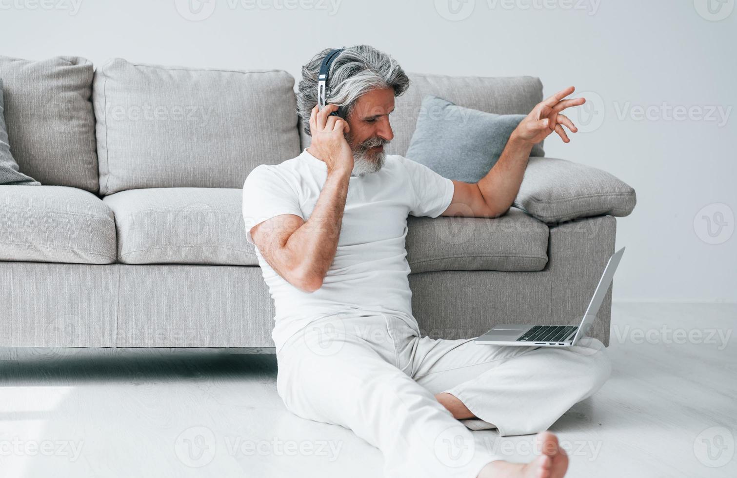 se sienta en el suelo cerca del sofá con una computadora portátil y escucha música. Senior hombre moderno con estilo con pelo gris y barba en el interior foto