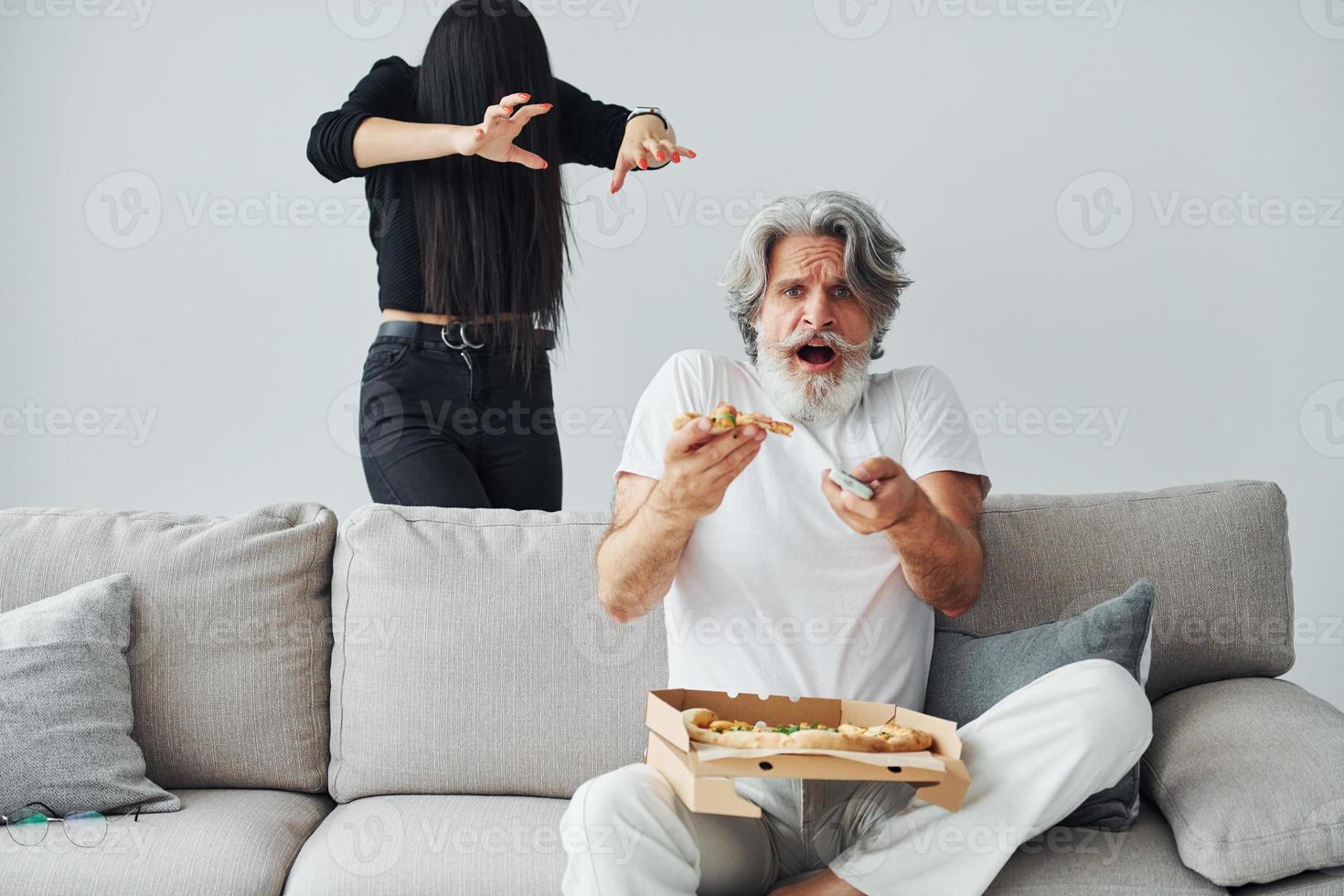 viendo la televisión con una mujer aterradora con el pelo negro cerca de él. Senior hombre moderno con estilo con pelo gris y barba en el interior foto