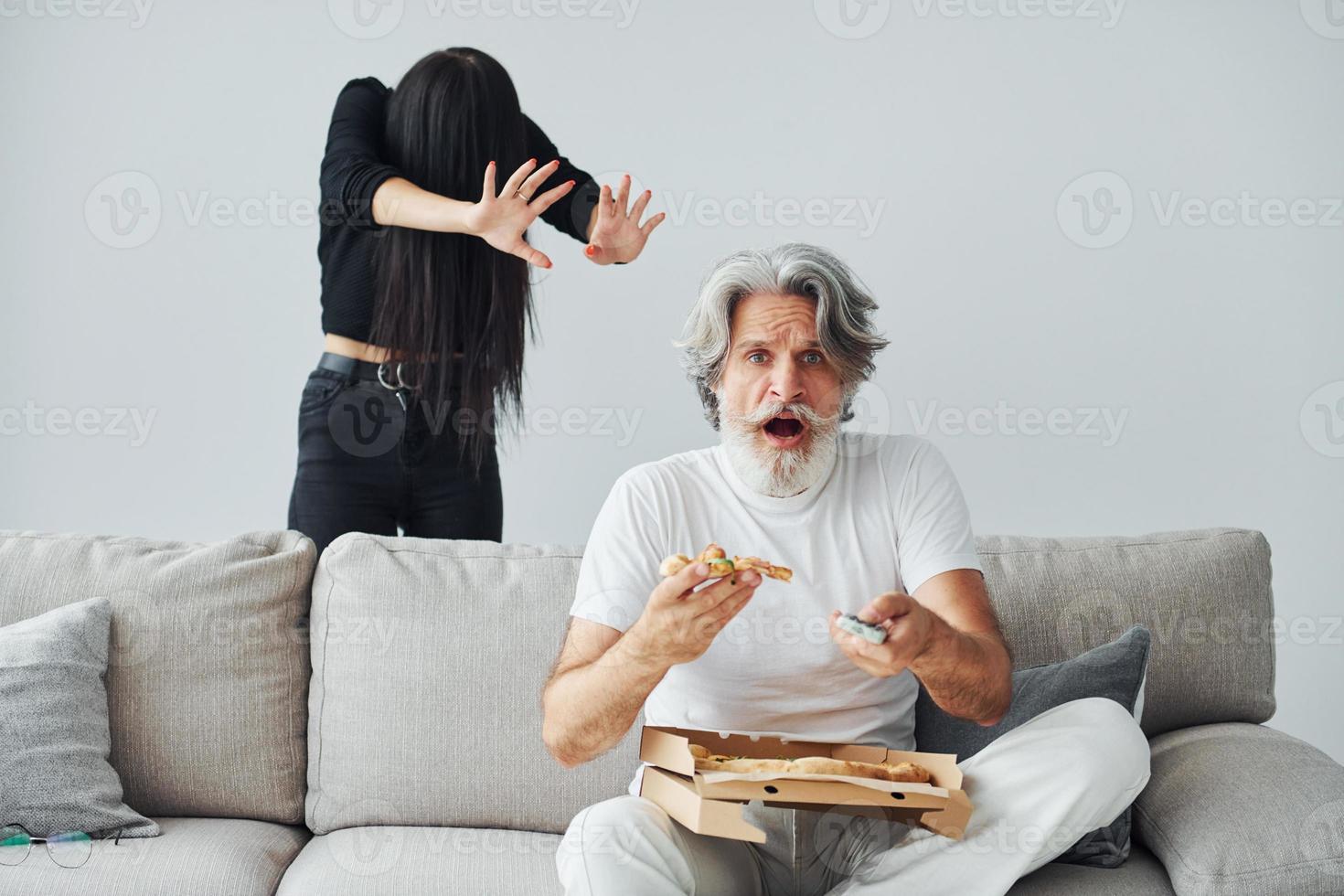 viendo la televisión con una mujer aterradora con el pelo negro cerca de él. Senior hombre moderno con estilo con pelo gris y barba en el interior foto