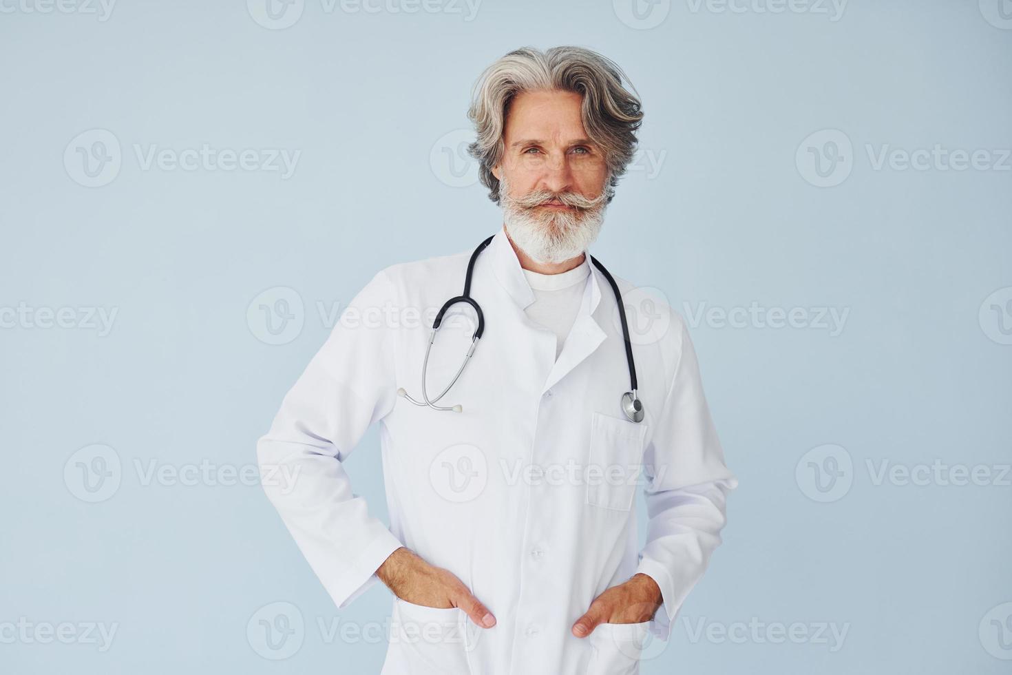 Doctor in white coat against wall. Senior stylish modern man with grey hair and beard indoors photo