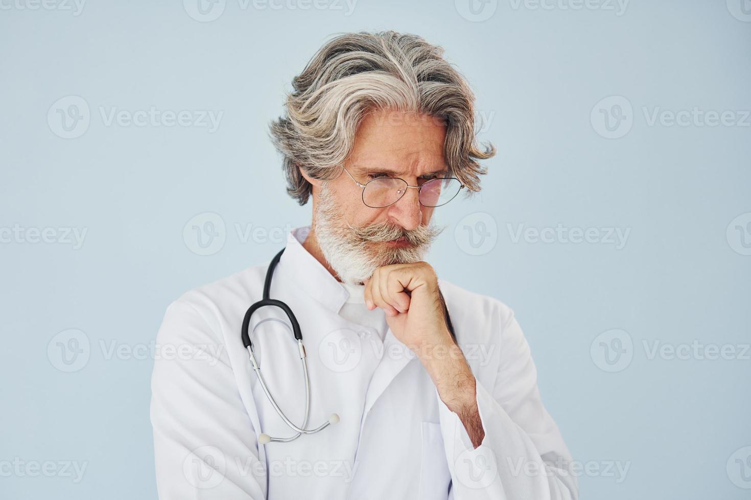 Doctor in white coat against wall. Senior stylish modern man with grey hair and beard indoors photo