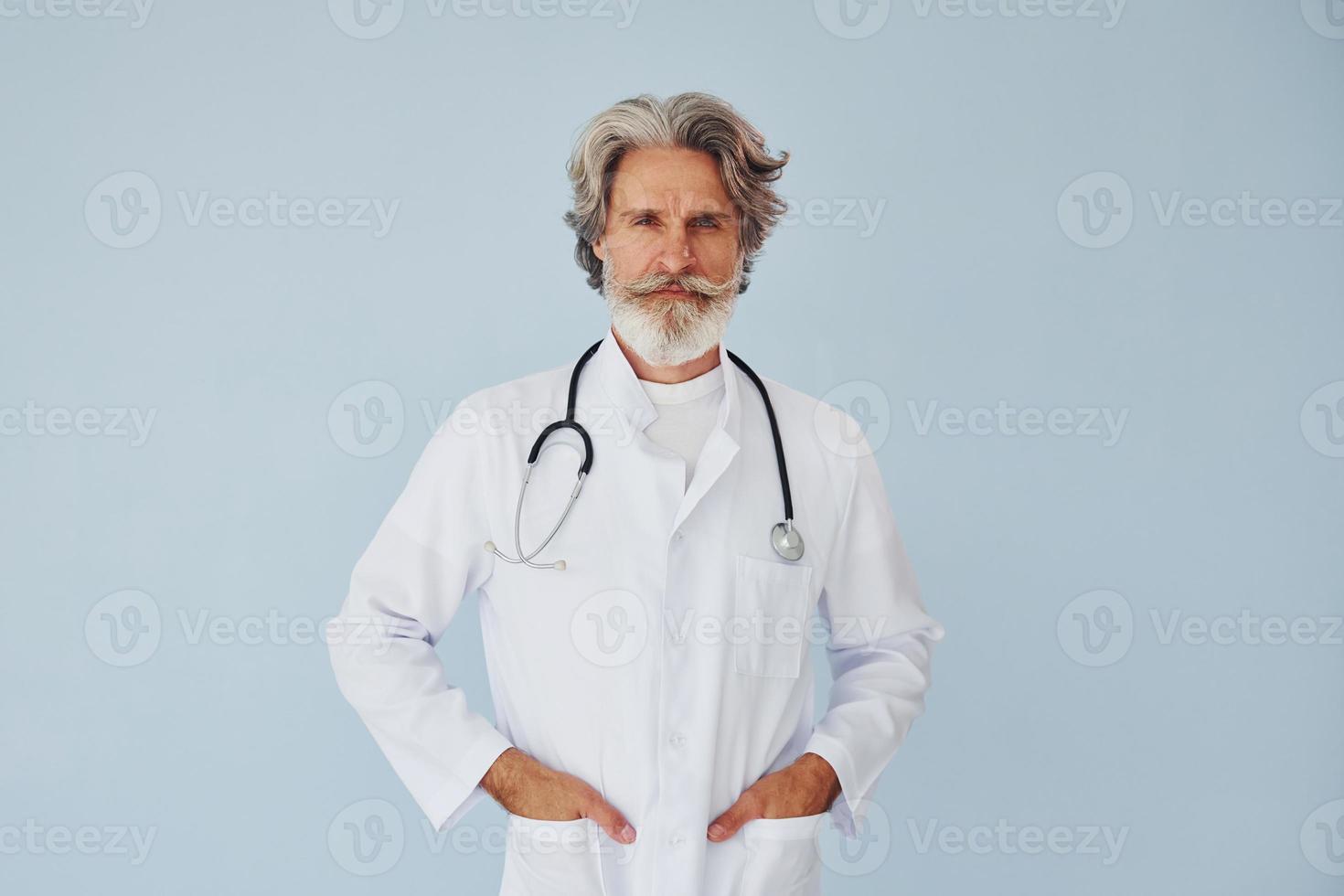 doctor en bata blanca contra la pared. Senior hombre moderno con estilo con pelo gris y barba en el interior foto