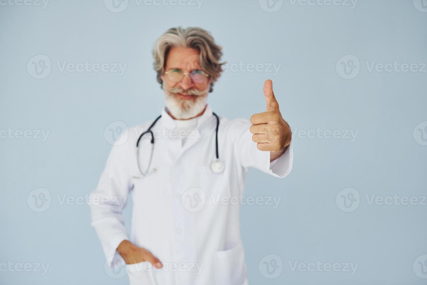 doctor en bata blanca contra la pared. Senior hombre moderno con estilo con pelo gris y barba en el interior foto