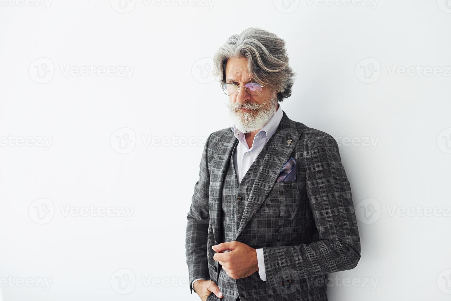 Posing for a camera while standing against white background. Senior stylish modern man with grey hair and beard indoors photo