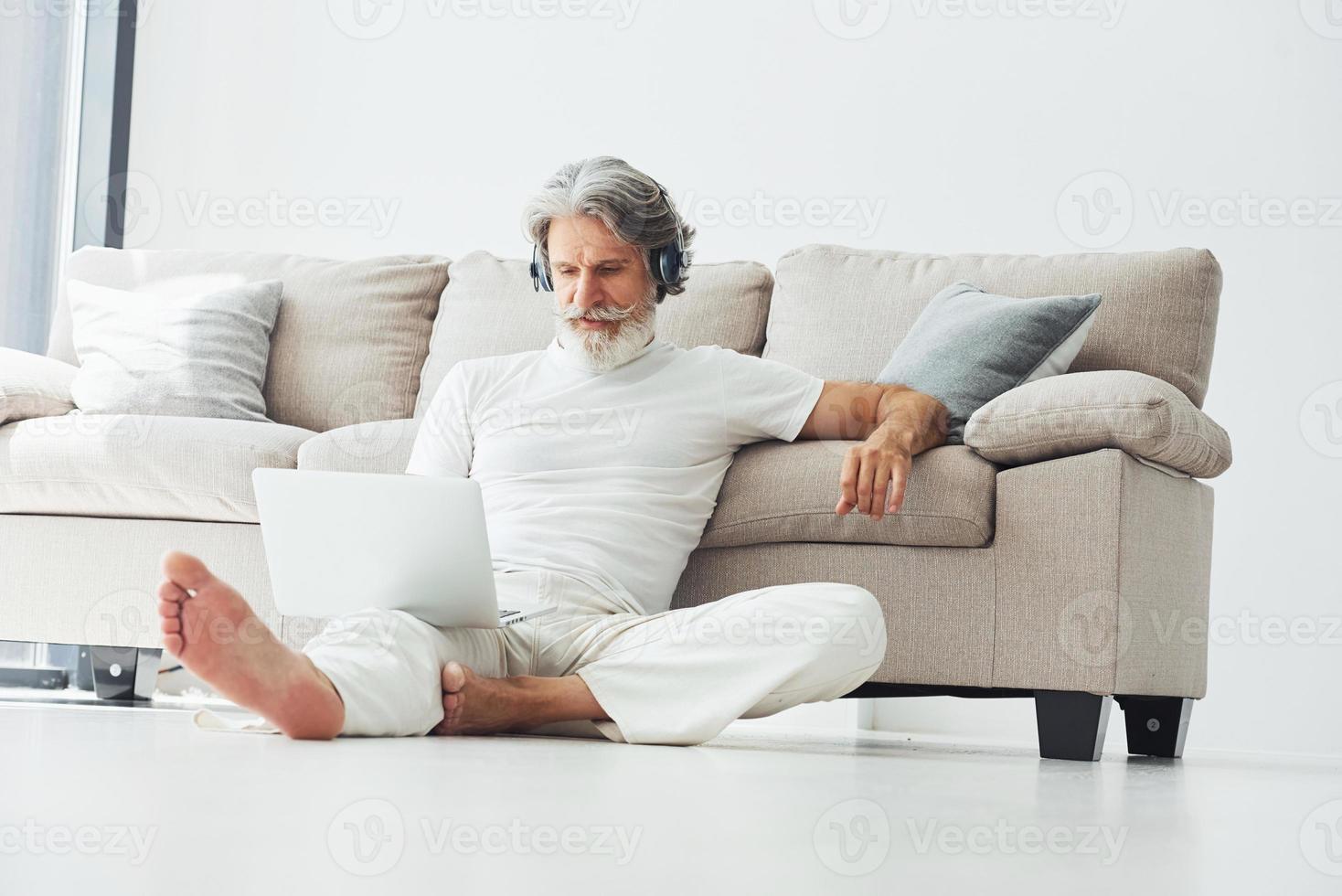 In headphones. Sits near sofa. Senior stylish modern man with grey hair and beard indoors photo