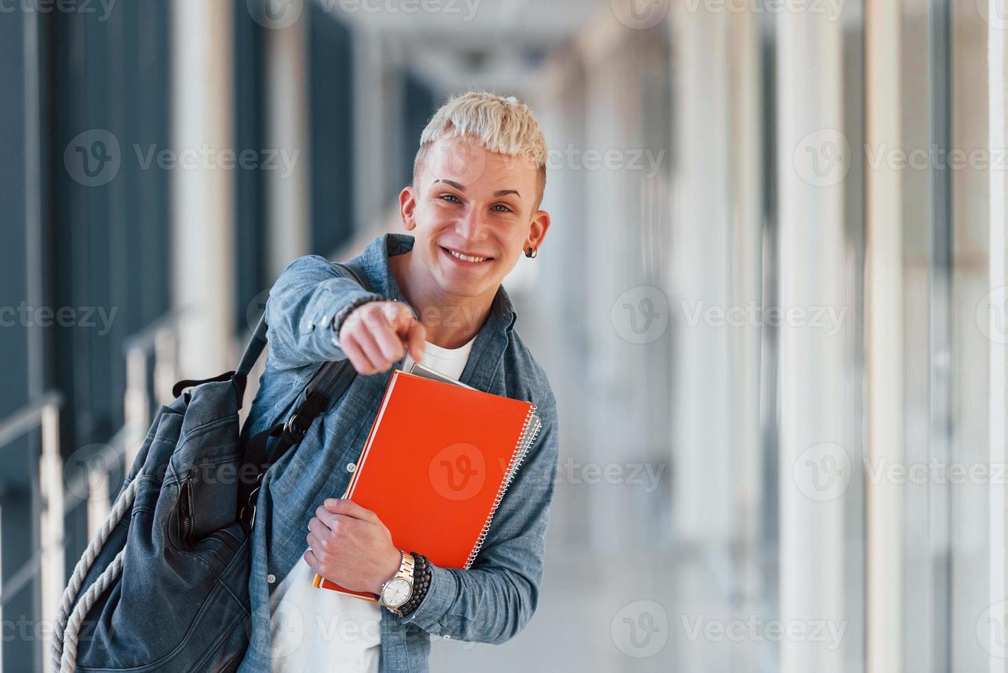 Male young student in jeans clothes is in corridor of a college with notepad in hands photo