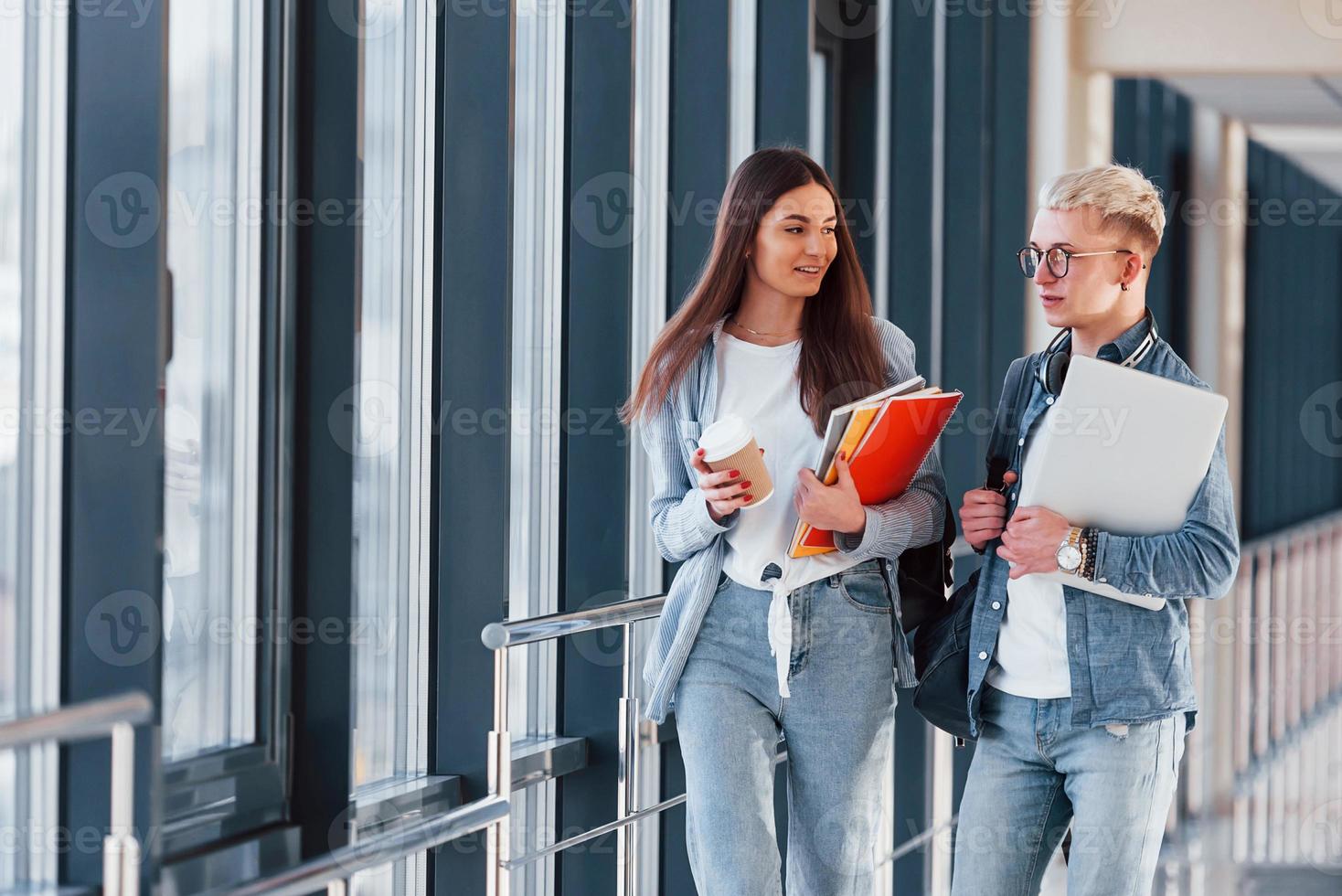 Two young student friends together in a corridor of a college walking together and holding notepad, laptop and cup of drink photo