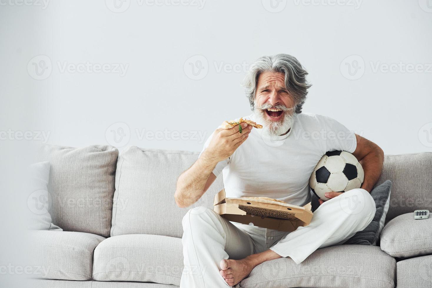 Soccer fan with pizza watches match. Senior stylish modern man with grey hair and beard indoors photo