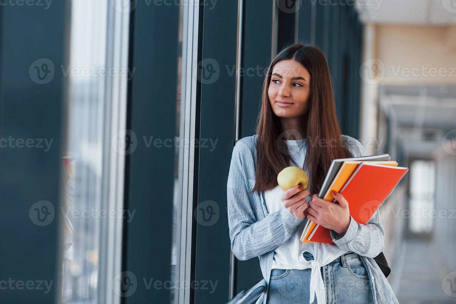 Female young student is in corridor of a college, holding notepads and an apple photo