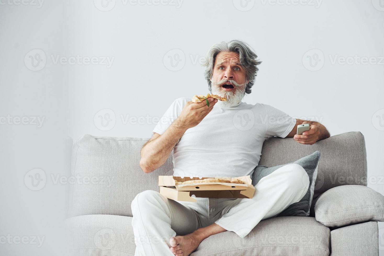 With delicious pizza. Watches TV show. Senior stylish modern man with grey hair and beard indoors photo