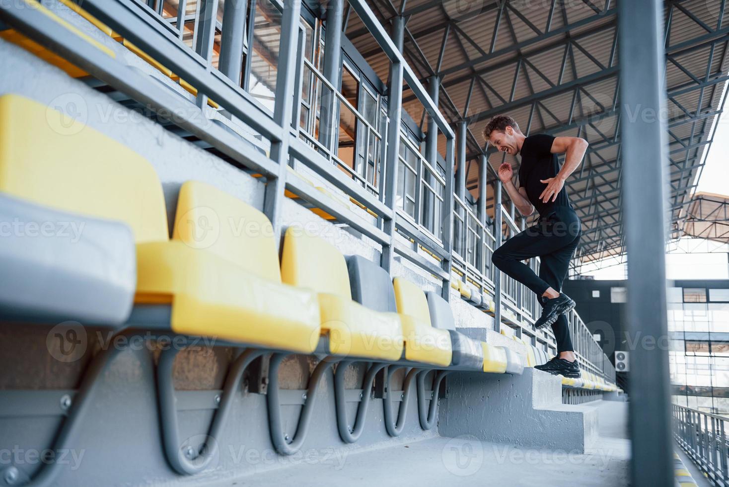 Running on the bleachers. Sportive young guy in black shirt and pants outdoors at daytime photo