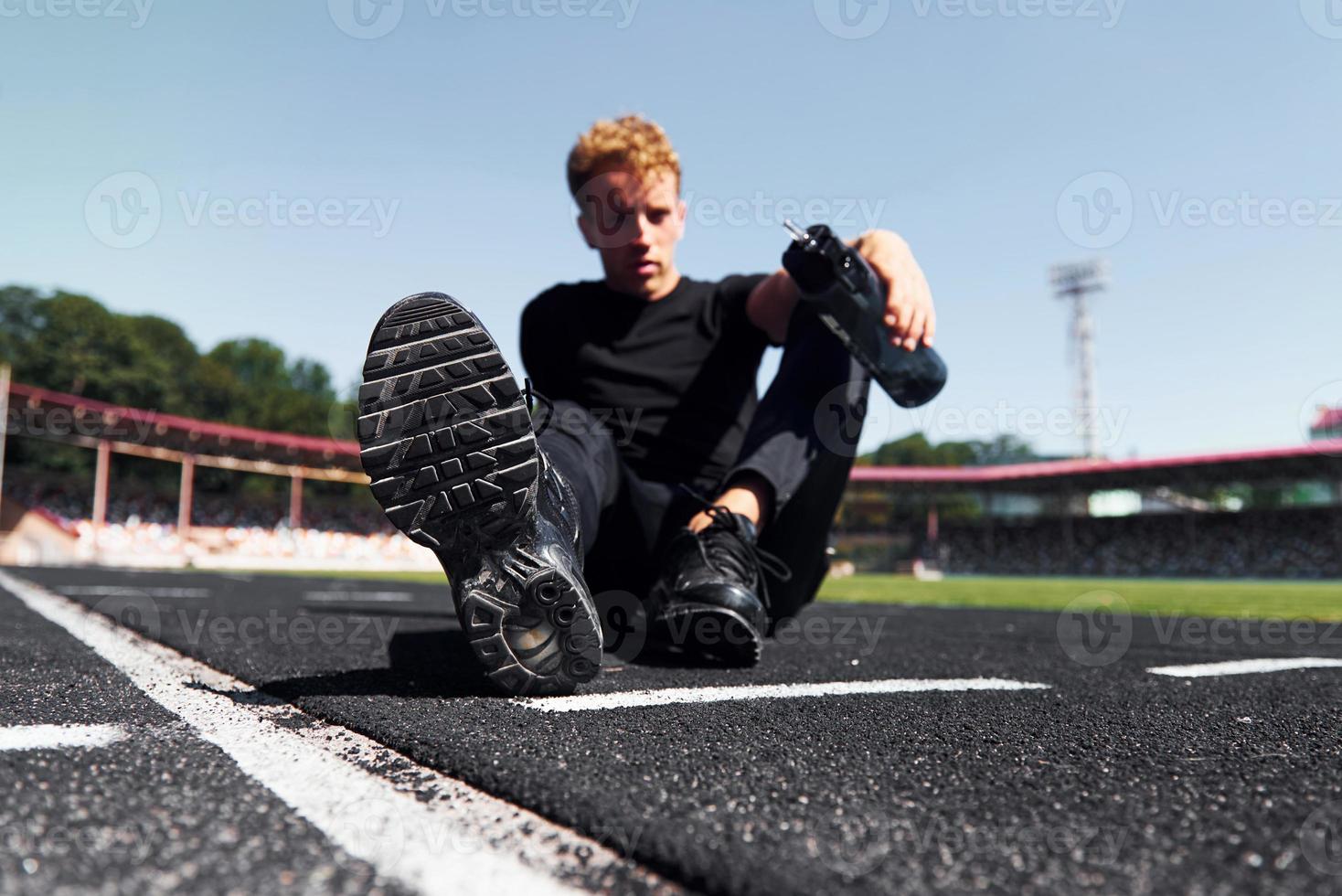 Tired runner sits on track and taking a break. Sportive young guy in black shirt and pants outdoors at daytime photo