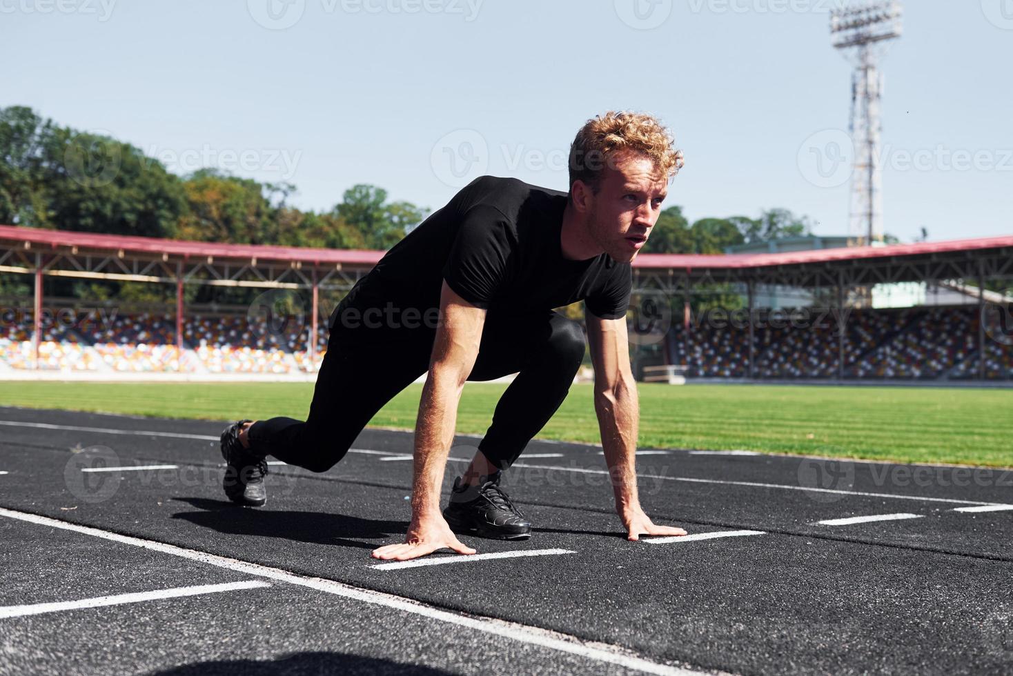 Runner is on starting position on track. Sportive young guy in black shirt and pants outdoors at daytime photo