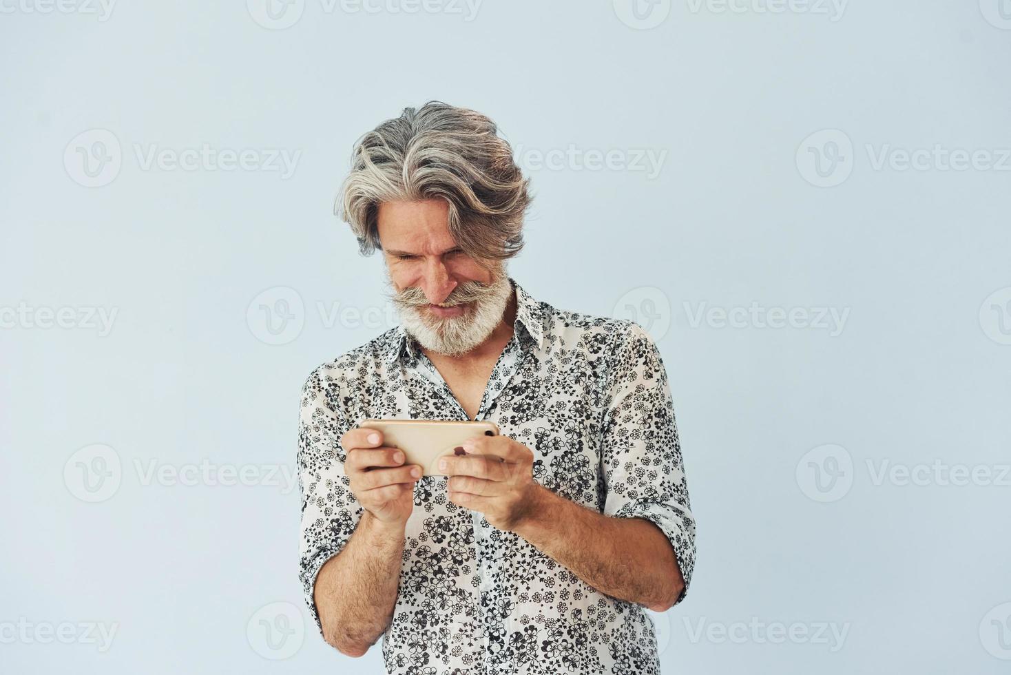 Senior stylish modern man with grey hair and beard indoors plays game by using phone photo