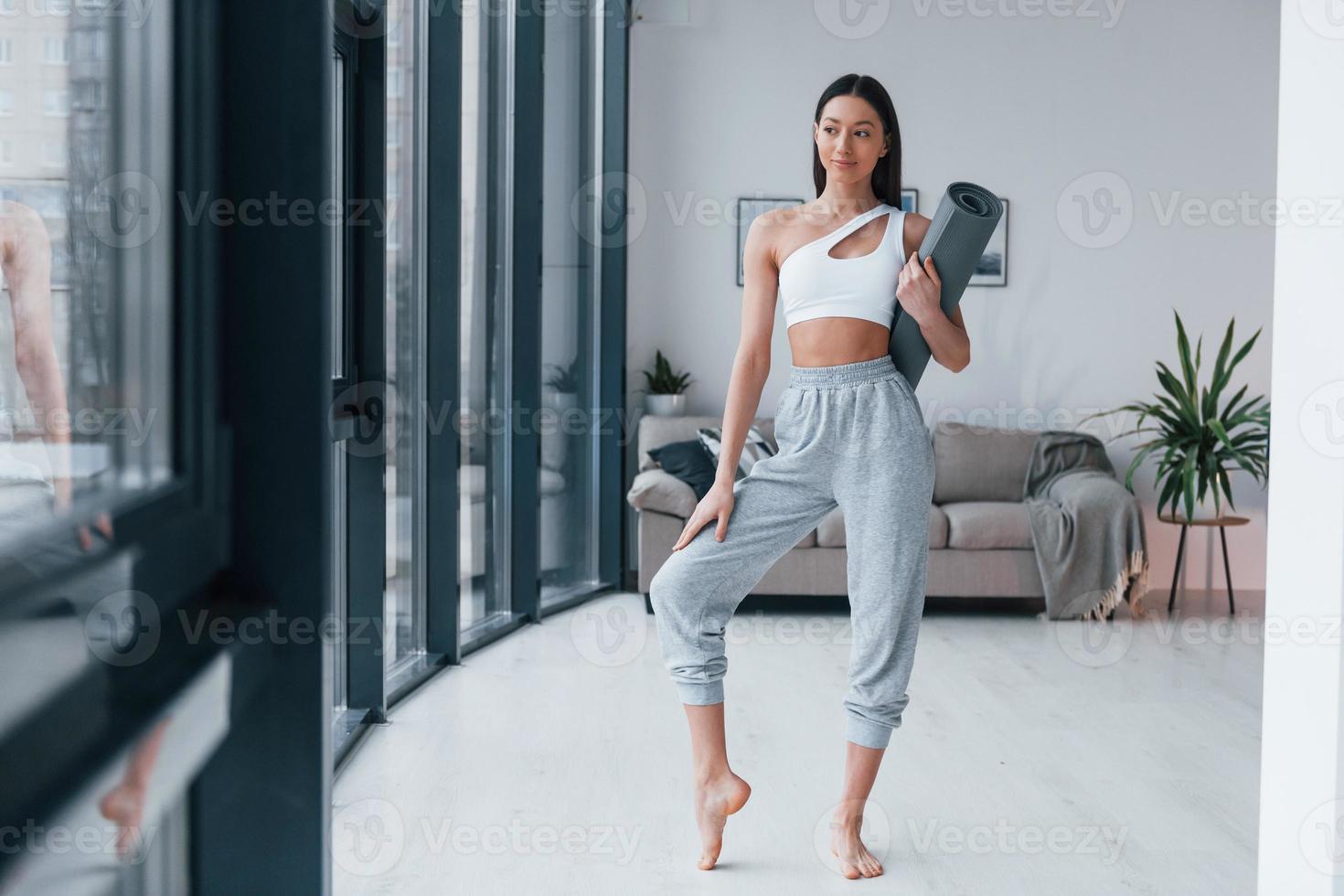 Standing and holds mat. Young woman with slim body shape in sportswear have fitness day indoors at home photo