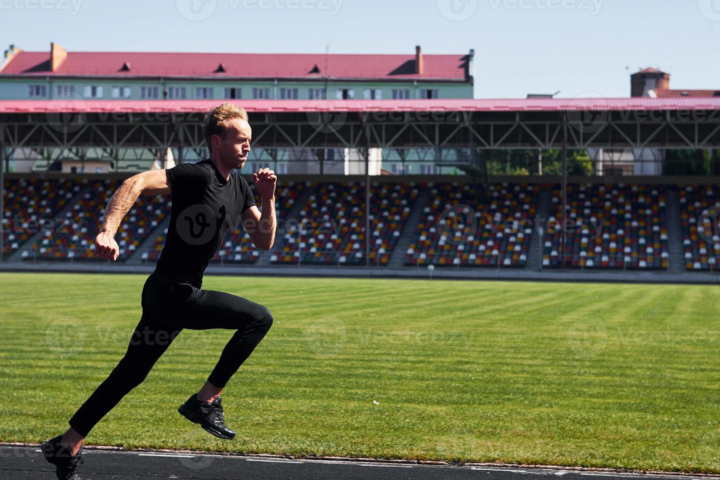 Side view of runner on field. Sportive young guy in black shirt and pants outdoors at daytime photo