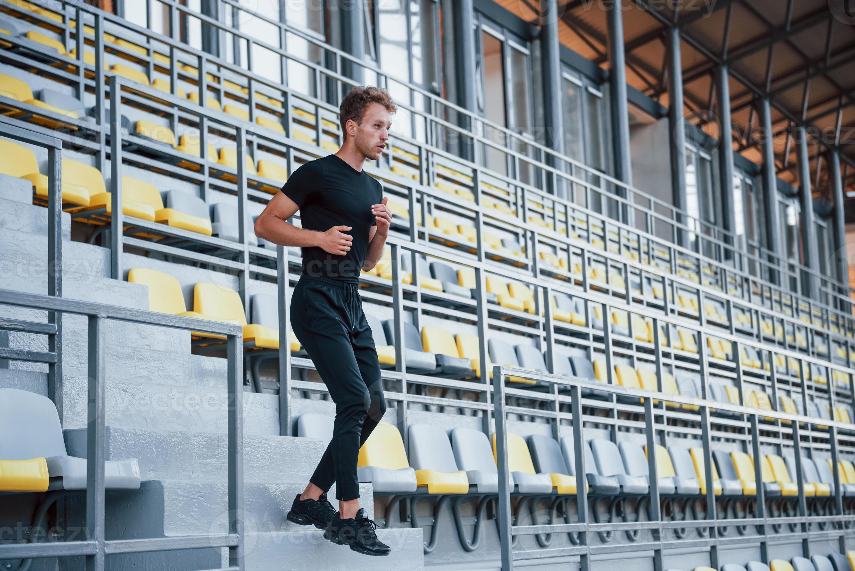 Running on the bleachers. Sportive young guy in black shirt and pants