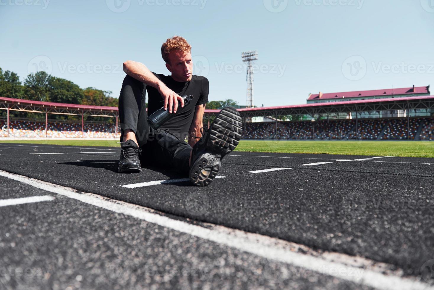 Tired runner sits on track and taking a break. Sportive young guy in black shirt and pants outdoors at daytime photo