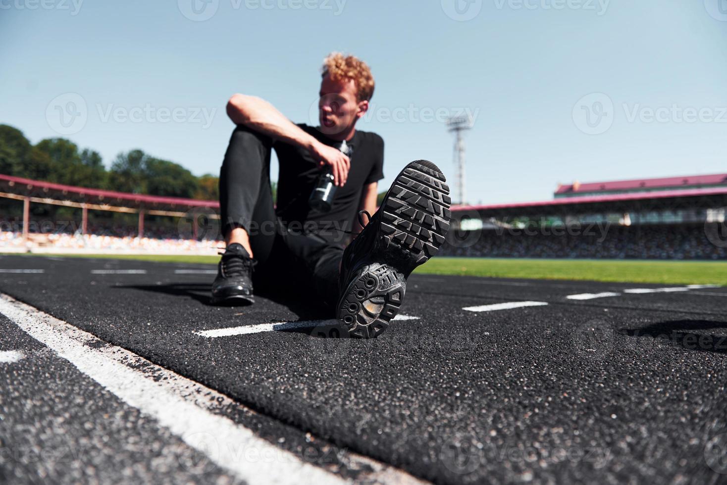 el corredor cansado se sienta en la pista y se toma un descanso. chico joven deportivo en camisa negra y pantalones al aire libre durante el día foto