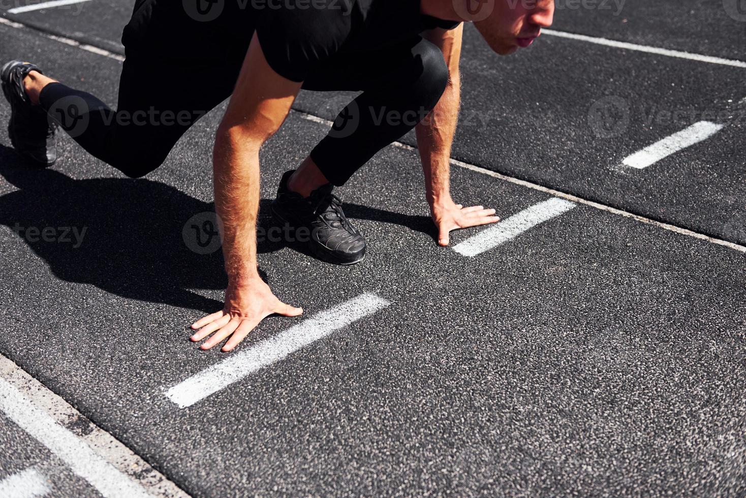Runner is on starting position on track. Sportive young guy in black shirt and pants outdoors at daytime photo