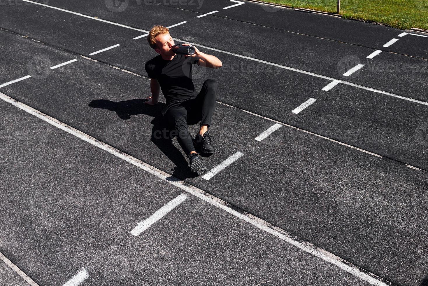 Tired runner sits on track and taking a break. Sportive young guy in black shirt and pants outdoors at daytime photo
