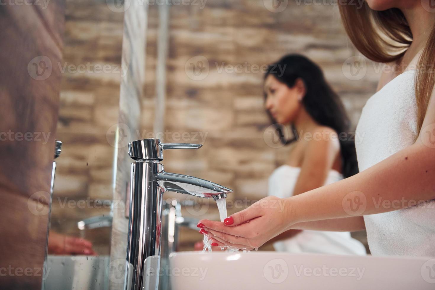 hermosa joven parada en el baño cerca del espejo y lavándose las manos. amigos detrás foto