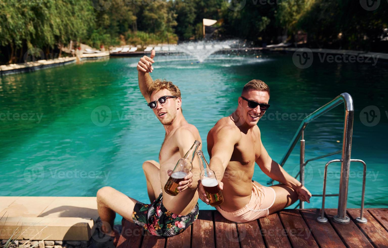 Two male friends sits near water outdoors with drinks in hands photo