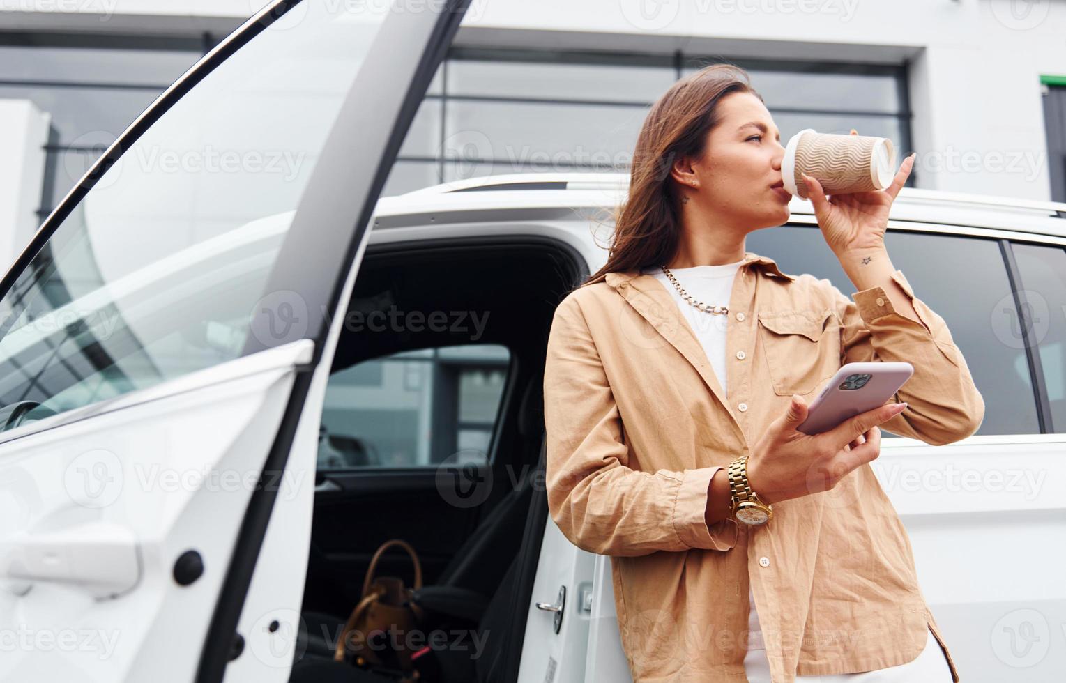 With coffee and phone. Fashionable beautiful young woman and her modern automobile photo
