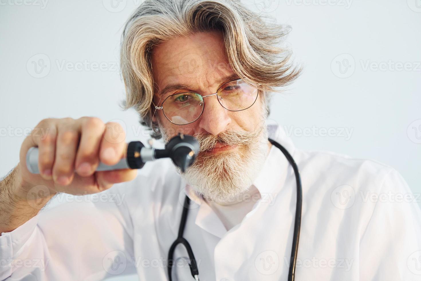 Portrait of senior male doctor with grey hair and beard in white coat is indoors in otorhinolaryngology clinic photo