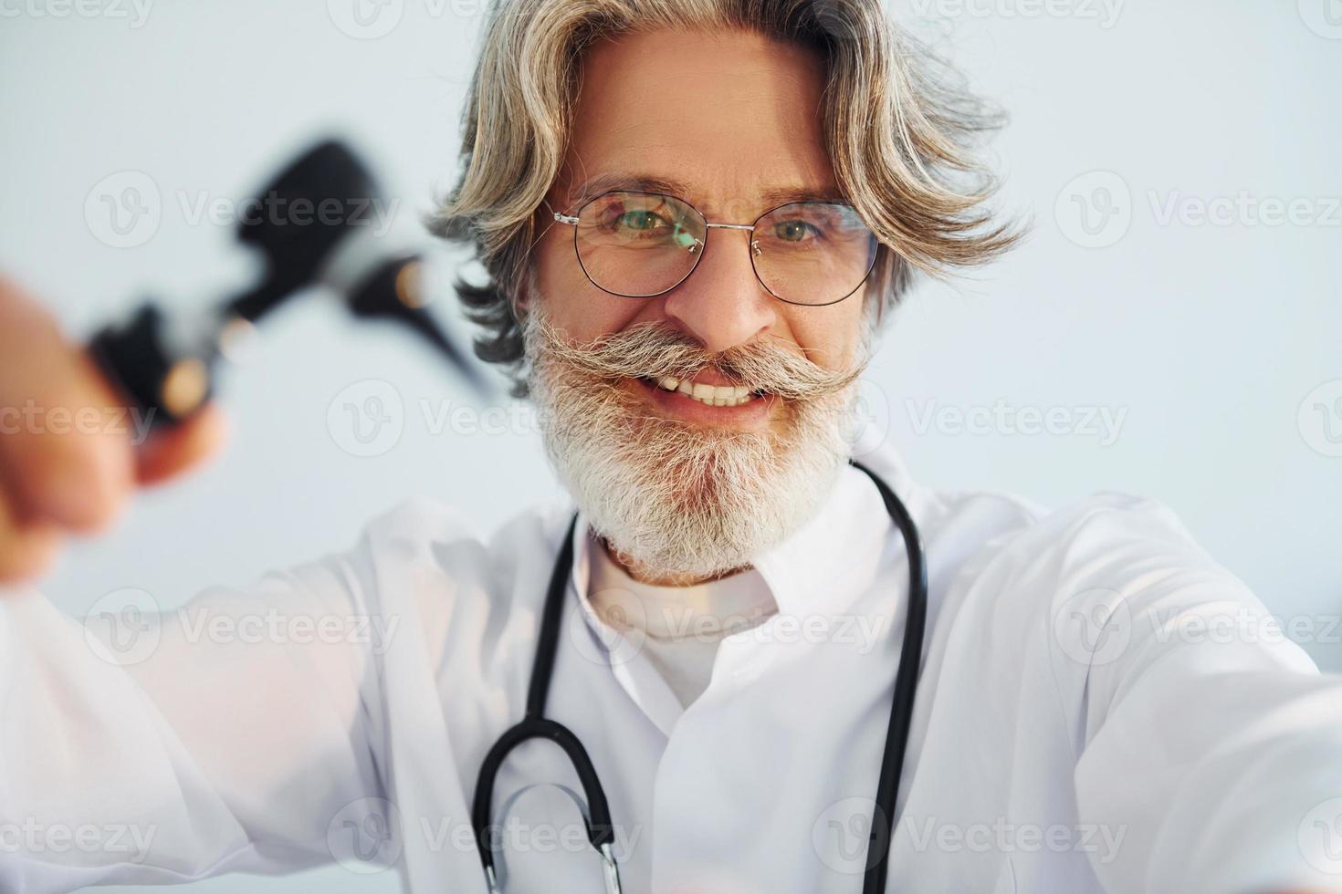 Portrait of senior male doctor with grey hair and beard in white coat is indoors in otorhinolaryngology clinic photo
