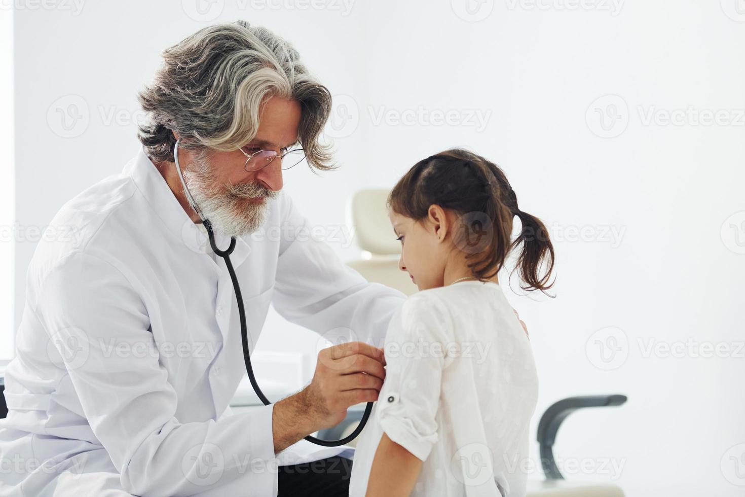 Senior male doctor with grey hair and beard in white coat measuring heart rate of little girl by stethoscope indoors in clinic photo