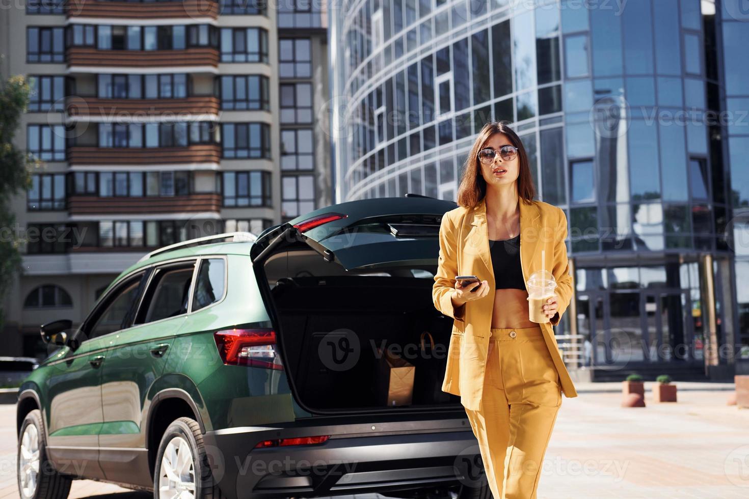 Modern business building at background. Young fashionable woman in burgundy colored coat at daytime with her car photo