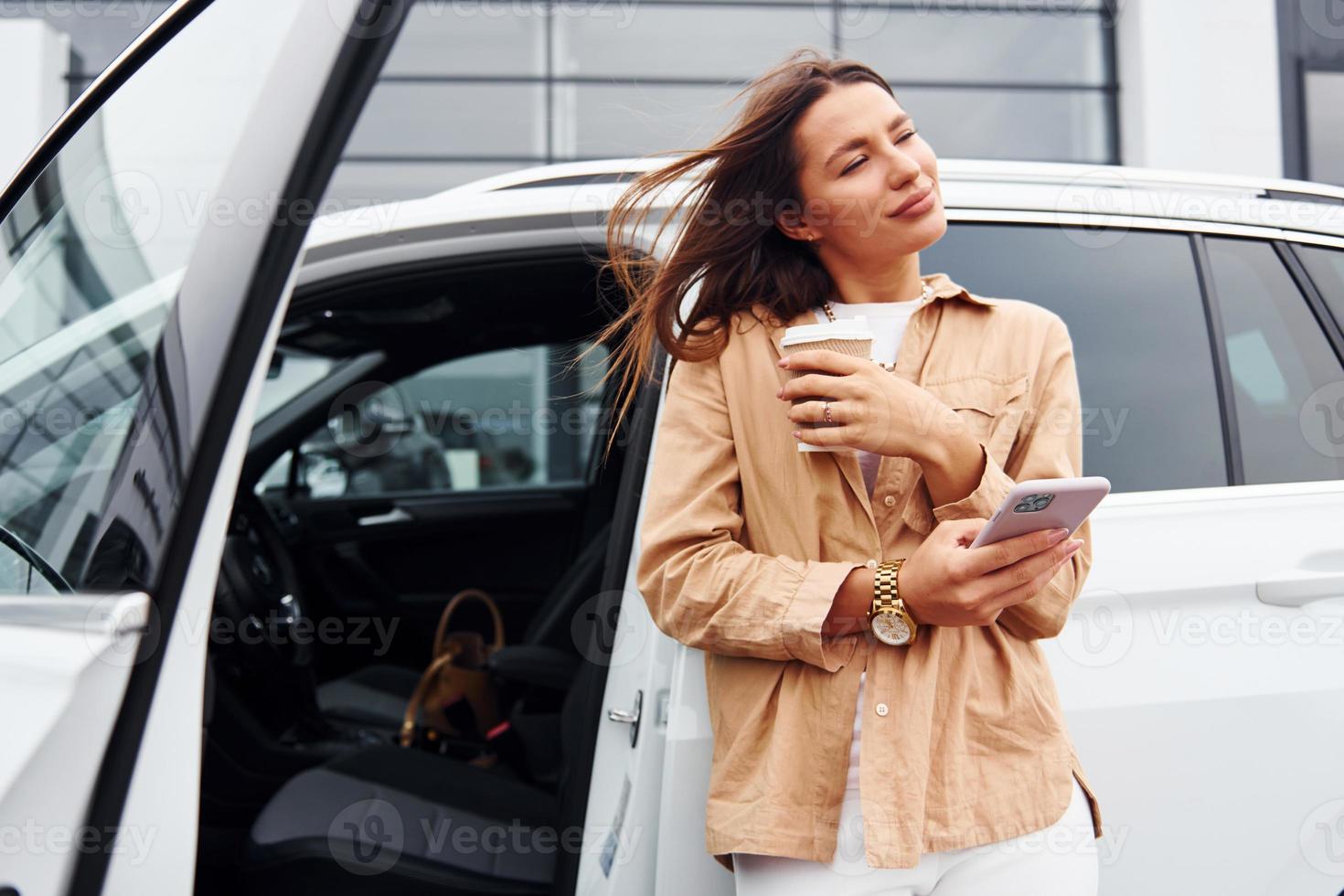 With coffee and phone. Fashionable beautiful young woman and her modern automobile photo