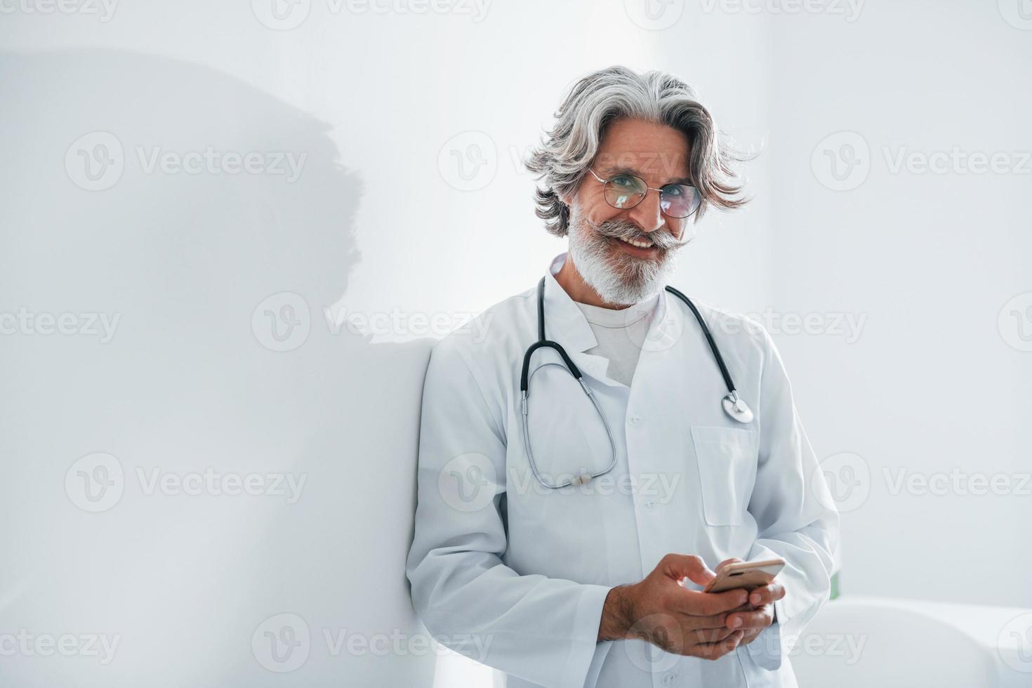 Senior male doctor with grey hair and beard in white coat is indoors in clinic photo