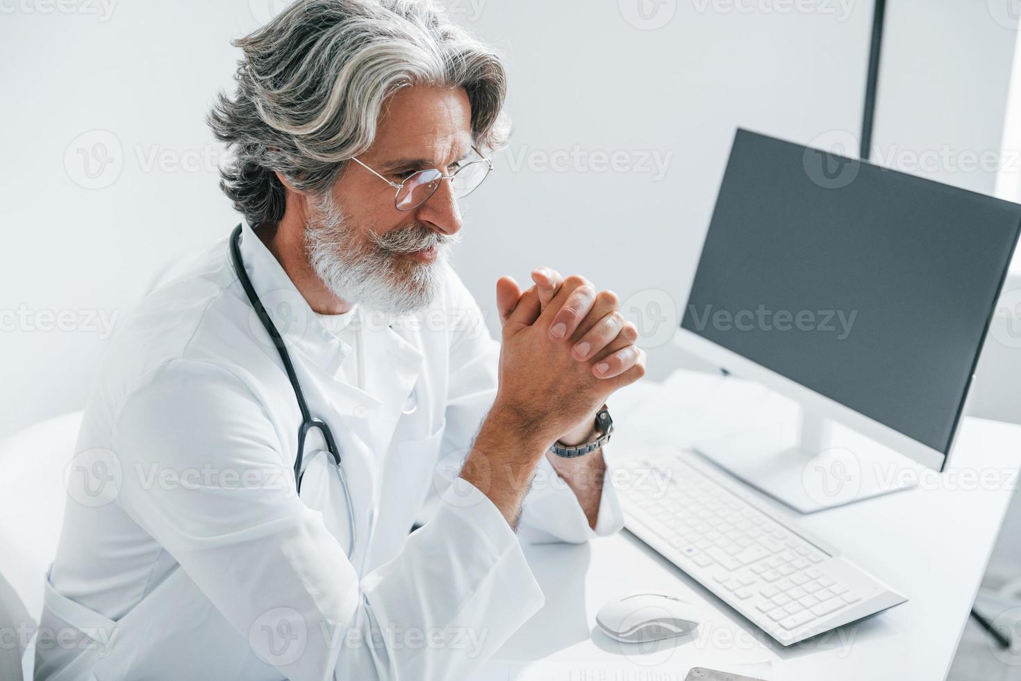 Portrait of senior male doctor with grey hair and beard in white coat is indoors in clinic photo