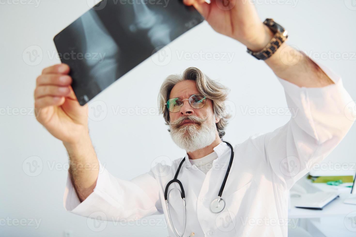 Looks at x-ray. Senior male doctor with grey hair and beard in white coat is indoors in clinic photo