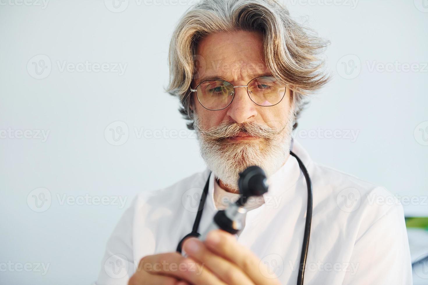 Portrait of senior male doctor with grey hair and beard in white coat is indoors in otorhinolaryngology clinic photo