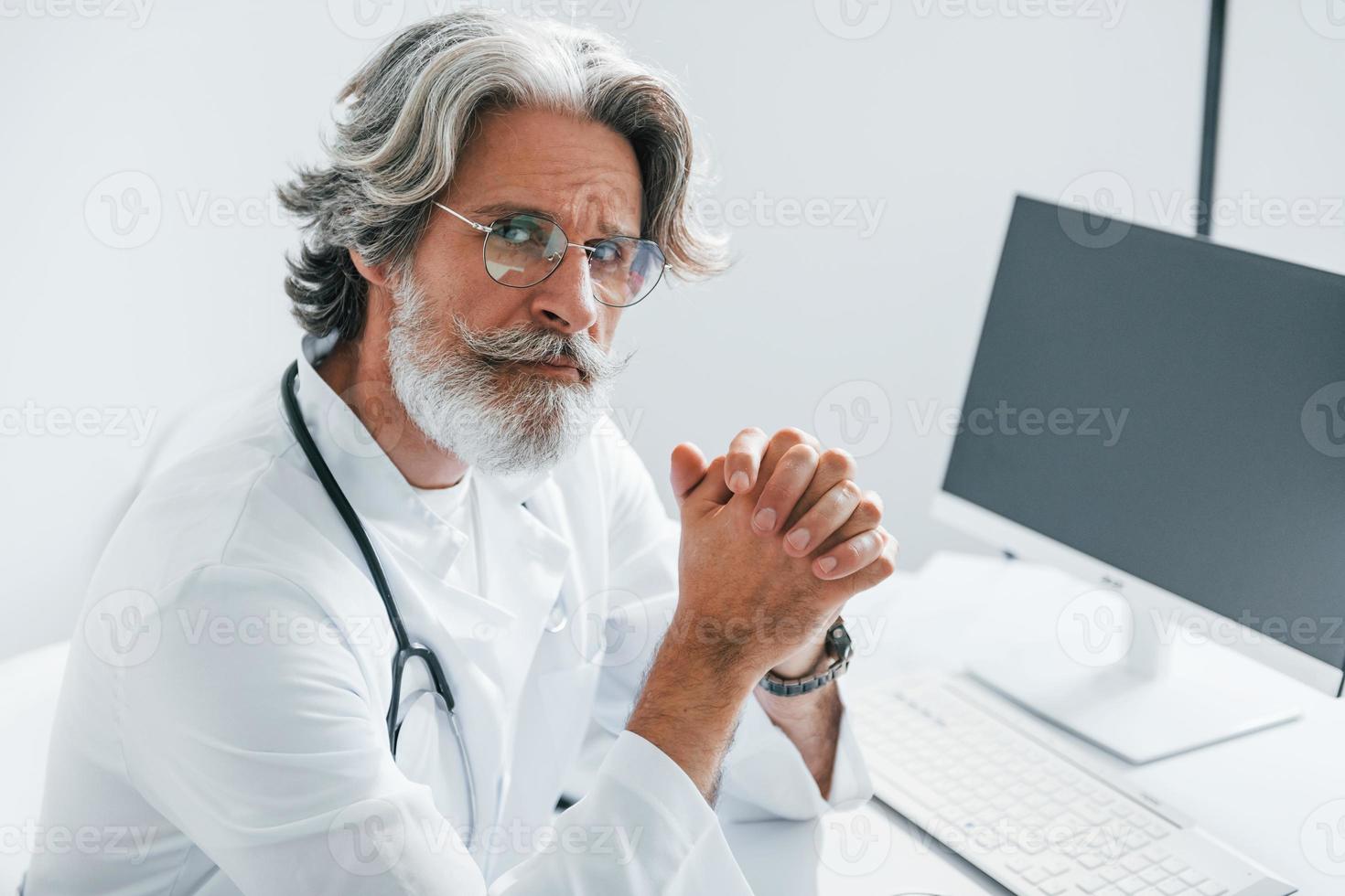 Portrait of senior male doctor with grey hair and beard in white coat is indoors in clinic photo