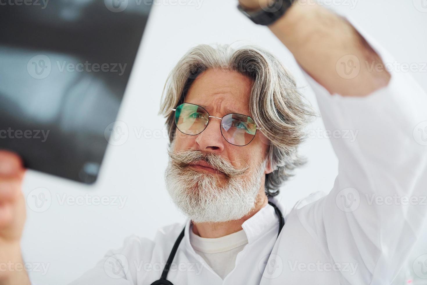 Looks at x-ray. Senior male doctor with grey hair and beard in white coat is indoors in clinic photo