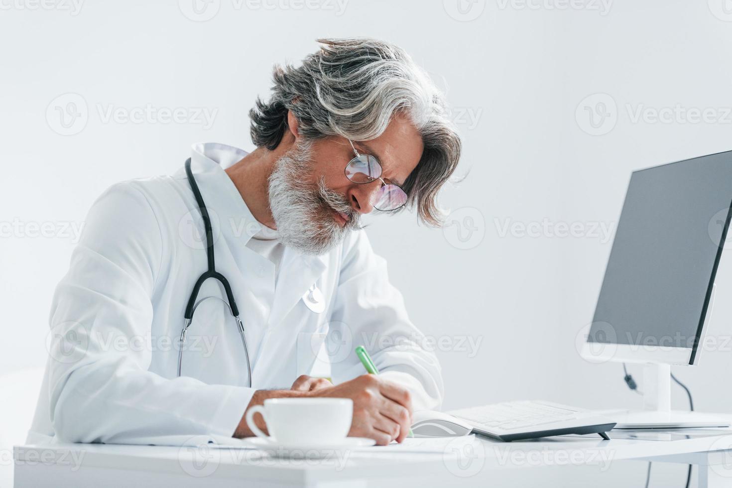 Writes by computer table. Senior male doctor with grey hair and beard in white coat is indoors in clinic photo