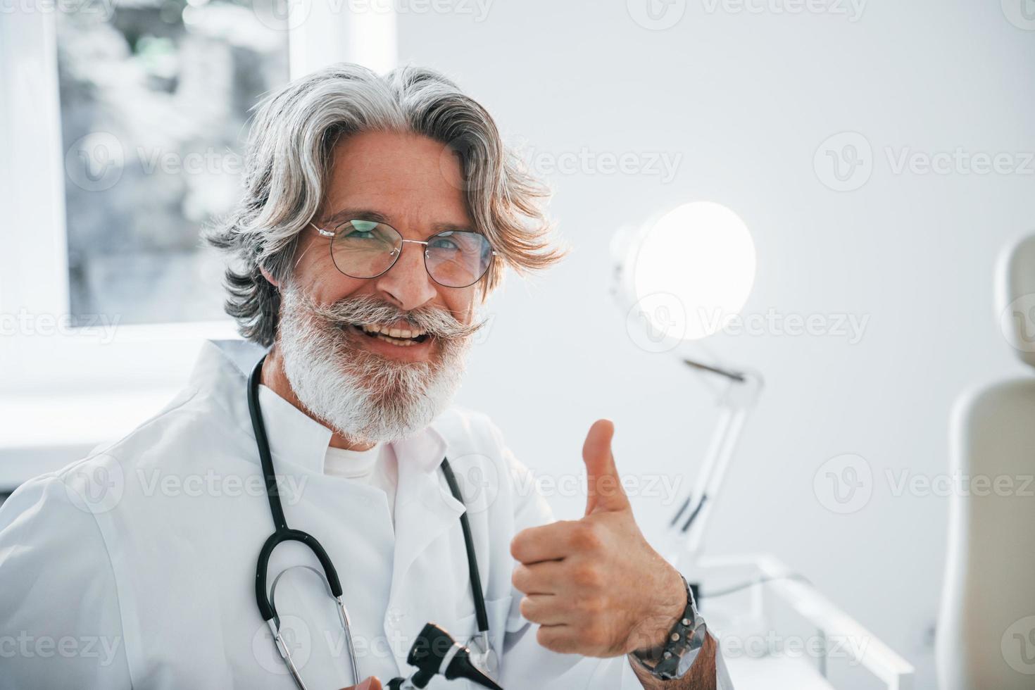 sonríe y muestra el pulgar hacia arriba. médico masculino senior con cabello gris y barba en bata blanca está en el interior de la clínica foto