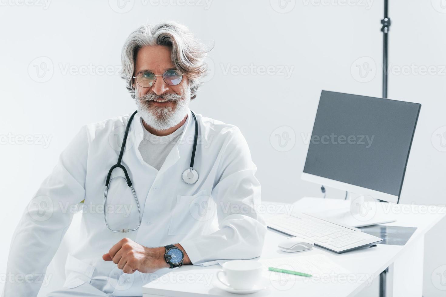 Works by computer table. Senior male doctor with grey hair and beard in white coat is indoors in clinic photo