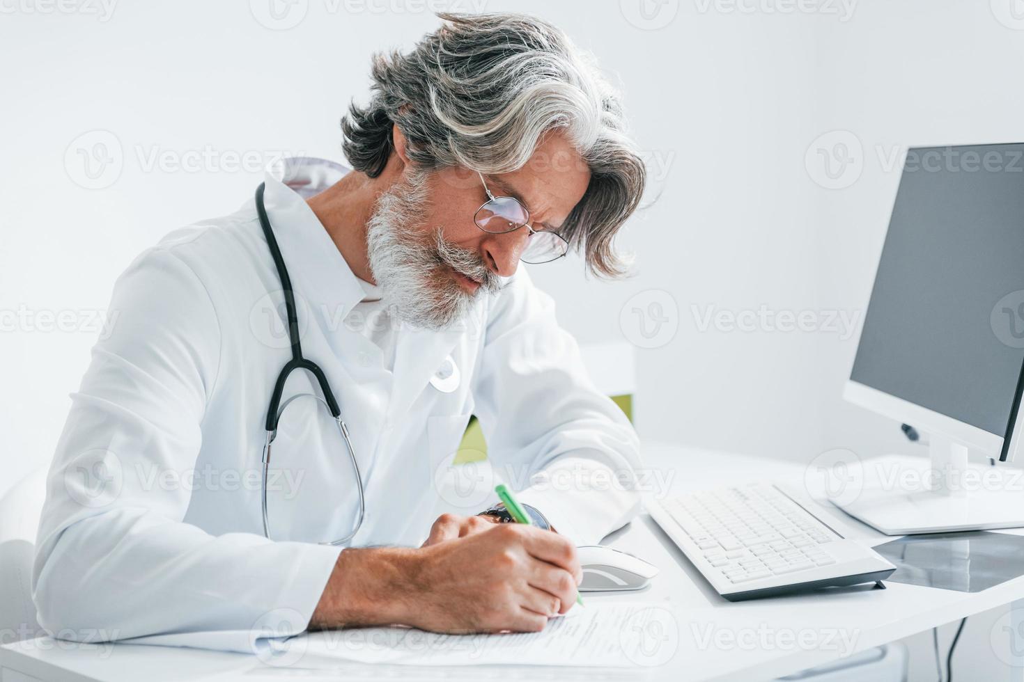 Writes by computer table. Senior male doctor with grey hair and beard in white coat is indoors in clinic photo