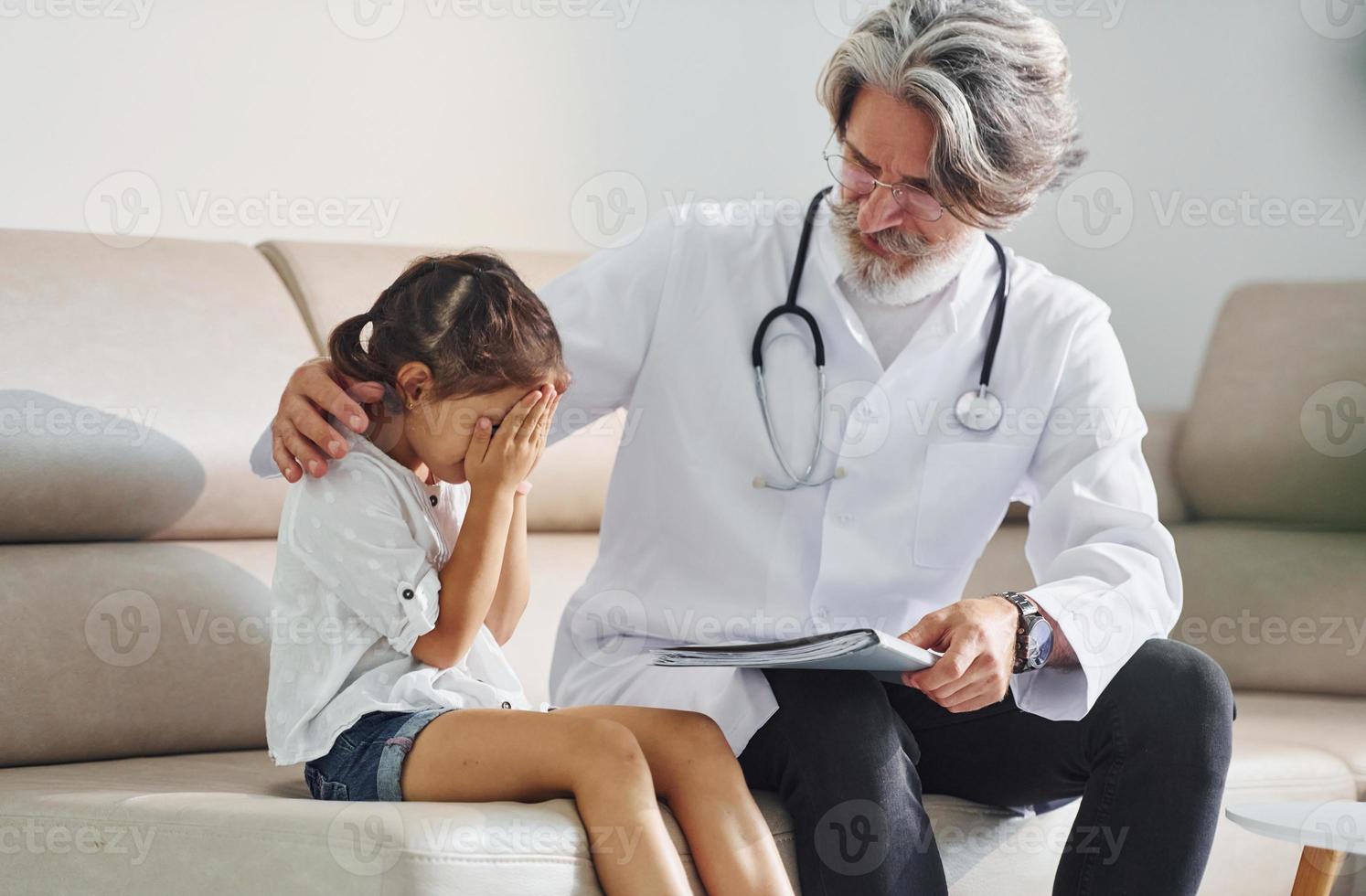 Talks with little sad girl. Working with customer. Senior male doctor with grey hair and beard in white coat is indoors in clinic photo