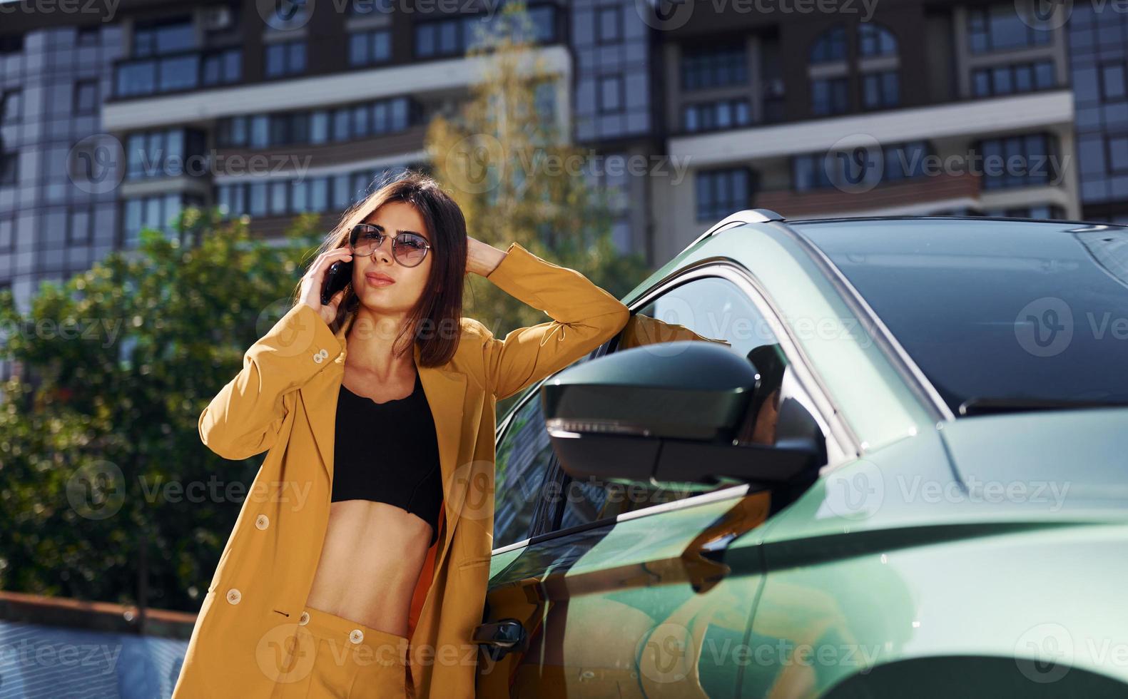 With smartphone. Young fashionable woman in burgundy colored coat at daytime with her car photo